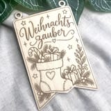 Ein rechteckiger Holzwimpel mit Gravur im Format A6 und zwei oberen Ösen liegt auf grauem Leinenstoff, mit dem Schriftzug Weihnachtszauber und Weihnachtsmotiven.