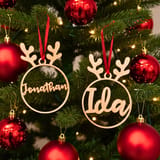Zwei personalisierte Christbaumkugeln in Rentier-Form mit den Namen Jonathan und Ida hängen an roten Bändern an einem grünen Tannenbaum zwischen roten Kugeln.