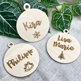 Könnte beinhalten: Drei kreisförmige, helle Holzanhänger mit einem kleinen Loch an der Oberseite. Jeder Anhänger ist mit einem lasergravierten Namen versehen: Kira mit einer Schneeflocke, Lisa Marie mit einem Tannenbaum und Philipp mit zwei Glocken.