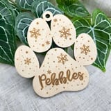 Könnte beinhalten: Ein helles Holzornament in Form eines Pfotenabdrucks mit drei ovalen Zehenballen und einem großen Hauptballen. Jeder Bereich ist mit Schneeflockenmustern und kleinen Punkten graviert. Der Name Pebbles ist in Schreibschrift eingraviert.
