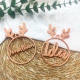 Zwei kupferfarbene, personalisierte Christbaumanhänger mit Rentiergeweih und den Namen Jonathan und Ida liegen nebeneinander auf einem Stroh- und Stoffuntergrund.