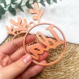 Eine Hand hält eine kupferfarbene, personalisierte Christbaumkugel mit dem Namen Ida und aufgesetztem Rentiergeweih vor einem Stroh- und Stoffhintergrund.