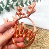 Eine Hand präsentiert nah eine kupferfarbene Christbaumkugel mit dem Namen Ida und Rentiergeweih, mit einem Ast im Hintergrund.