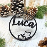 Ein schwarzer 3D-gedruckter Weihnachtsanhänger mit dem Namen Luca und Sternen, umgeben von Tannenzapfen und Tannengrün.