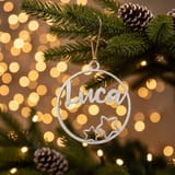 Ein silberner Weihnachtsanhänger mit dem Namen Luca hängt an einem Tannenbaum vor warmem Lichterbokeh.