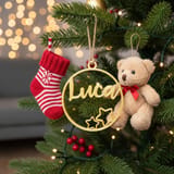 Ein goldener runder 3D-Druck-Weihnachtsanhänger mit dem Namen Luca und zwei Sternen hängt an einem Weihnachtsbaum neben einem Plüschbären und einem rot-weißen Strumpf.