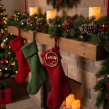Ein goldener runder Weihnachtsanhänger mit dem Namen Luca hängt an einem roten Weihnachtssocken, der an einem Kamin mit Tannengrün und Kerzen befestigt ist.