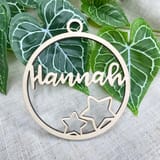 Könnte beinhalten: Ein kreisförmiges Ornament aus hellem Holz mit dem Namen Hannah in Schreibschrift, ergänzt durch zwei ausgeschnittene Sternformen unter dem Text und eine kleine kreisförmige Öse an der Oberseite zum Aufhängen.