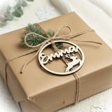 Könnte beinhalten: Ein quadratisches Geschenkpaket in braunem Packpapier, umwickelt mit dünner Jutekordel. Ein runder Holzanhänger ist an der Kordel befestigt, der den Namen Emma in Schreibschrift sowie die ausgeschnittene Silhouette eines Rentiers zeigt.