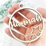 Könnte beinhalten: Ein kreisförmiges Ornament aus hellem Holz mit dem Namen Hannah in Schreibschrift. Das Design enthält zwei sternförmige Ausschnitte unten rechts und ein kleines kreisförmiges Loch oben zum Aufhängen, mit dunklen, lasergeschnittenen Kanten.