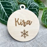 Könnte beinhalten: Ein kreisförmiges Ornament aus hellem Holz mit einem kleinen Loch an der Oberseite zum Aufhängen. Der Name Kira ist in Schreibschrift in die Mitte graviert, mit einem detaillierten Schneeflockenmotiv direkt unter dem Namen auf der Holzoberfläche.