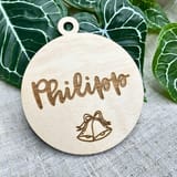 Könnte beinhalten: Ein kreisförmiges Ornament aus hellem Holz mit einem kleinen Loch an der Oberseite zum Aufhängen. Die Oberfläche ist mit dem Namen Philipp in Schreibschrift lasergraviert, unter dem sich eine einfache Strichzeichnung zweier kleiner Glocken befindet.
