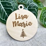 Könnte beinhalten: Ein kreisförmiges Ornament aus hellem Holz mit einem kleinen Loch an der Oberseite zum Aufhängen. Der Text Lisa Marie ist in einer Schreibschrift mittig eingraviert, mit einem einfachen stilisierten Weihnachtsbaum-Symbol unter dem Namen.