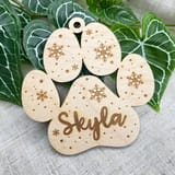 Könnte beinhalten: Ein helles Holzornament in Form eines Pfotenabdrucks mit eingravierten Schneeflockenmustern und kleinen Punkten. Der Name Skyla ist in Schreibschrift in den mittleren Ballen der Pfote lasergraviert, mit einem kleinen Loch zum Aufhängen.