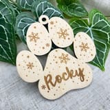 Könnte beinhalten: Ein helles Holzornament in Form eines Pfotenabdrucks mit dem eingravierten Namen Rocky in Schreibschrift. Die Oberfläche ist mit lasergeschnittenen Schneeflockenmustern und kleinen Punkten verziert und hat ein Loch zum Aufhängen.