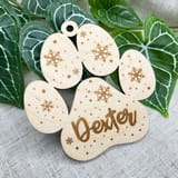 Könnte beinhalten: Ein helles Holzornament in Form eines Pfotenabdrucks mit einem kleinen runden Loch an der Oberseite zum Aufhängen. Die Oberfläche ist mit Schneeflockenmustern, kleinen Punkten und dem Namen Dexter in Schreibschrift graviert.
