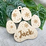 Könnte beinhalten: Ein helles Holzornament in Form eines Pfotenabdrucks mit lasergravierten Schneeflockenmustern und kleinen Punkten. Der Name Jack ist in Schreibschrift auf dem mittleren Ballen des Pfotenabdrucks eingraviert, mit einem Loch oben.