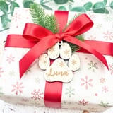 Könnte beinhalten: Ein quadratisches Geschenkpaket in weißem Papier mit rotem und grünem Schneeflockenmuster, verziert mit einem roten Satinband und einer Schleife. Ein heller Holzanhänger in Pfotenform mit der Gravur Luna und Schneeflocken ist befestigt.