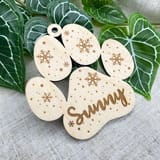 Könnte beinhalten: Ein helles Holzornament in Form eines Pfotenabdrucks mit eingravierten Schneeflockenmustern und verstreuten Punkten. Der Name Sunny ist in Schreibschrift auf dem mittleren Ballen eingraviert, mit einem kleinen Loch an der Oberseite.