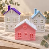 Drei gedruckte Winterhäuser in Weiß und Rosa mit violettem, hellblauem und weißem Dach auf hellem Leinen, mit Eukalyptus und warmen Lichtern im Hintergrund