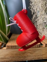 Ein leuchtend rotes, 3D-gedrucktes Schlittschuh-Ornament mit glatter Oberfläche und einem weißen Band zum Aufhängen an der Oberseite. Der Schlittschuh hat weiße Schnürsenkel, die vorne zu einer Schleife gebunden sind, und eine rote Kufe.