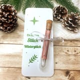 Eine weiße rechteckige Karte mit einem grünen Stern, einem Blattsymbol und dem grünen Text "Ein kleines Stück Winterglück". Ein durchsichtiges Glasreagenzglas mit Korken und einem gerollten roten Papier ist mit einem silbernen Band befestigt.