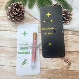 Eine weiße rechteckige Karte mit grünem Text "Ein kleines Stück Winterglück" und ein kleines Glasröhrchen mit Korken, neben einer schwarzen rechteckigen Karte mit goldenem Text "Sternstunden für dich".