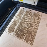 Drei gravierte winterliche Lesezeichen auf einer Holzplatte im Laser – quadratische, KI-bearbeitete Fotofassung.