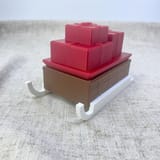 Originalfoto von Ninas gedrucktem Geschenkschlitten mit braunem Geheimfach, roten Geschenkpaketen und weißen Kufen, schräg auf hellem Stoff.