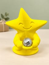 KI-bearbeitetes Produktbild: Gelbe Tanzfunke-Sternfigur mit separater silberner Mini-Discokugel vor salbeigrünem Hintergrund und einer Geschenkverpackung.