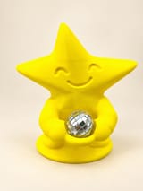 KI-bearbeitetes Produktbild: Gelbe Tanzfunke-Sternfigur mit lächelndem Gesicht, integrierter Schale und separater silberner Mini-Discokugel vor cremefarbenem Studiohintergrund.