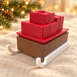 Geschenkschlitten mit braunem Geheimfach, rotem Geschenkdeckel und weißen Kufen auf cremefarbenem Leinen vor warmen Weihnachtslichtern und Tannengrün.
