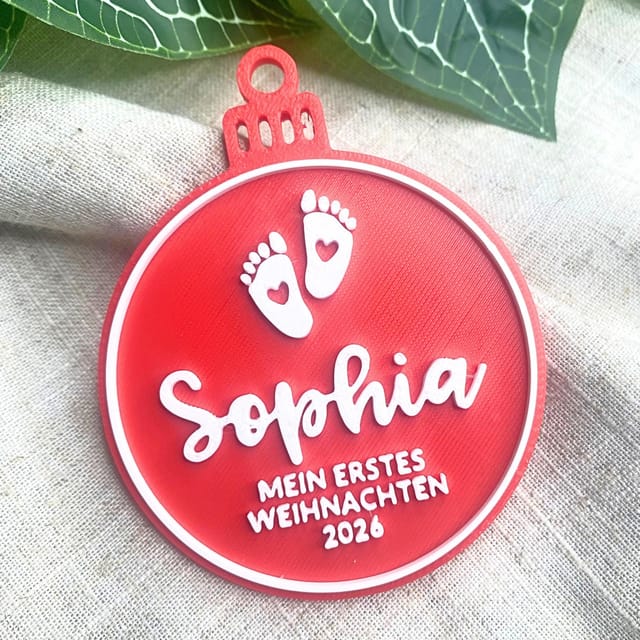 Eine rote 3D-gedruckte Christbaumkugel mit weißer Aufschrift Sophia, Babyfüßen und der Inschrift Mein erstes Weihnachten 2026.