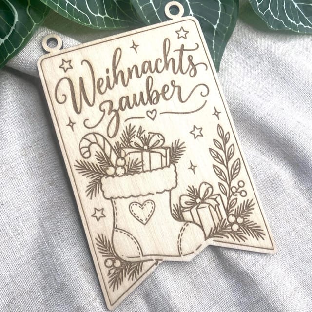 Ein rechteckiger Holzwimpel mit Gravur im Format A6 und zwei oberen Ösen liegt auf grauem Leinenstoff, mit dem Schriftzug Weihnachtszauber und Weihnachtsmotiven.