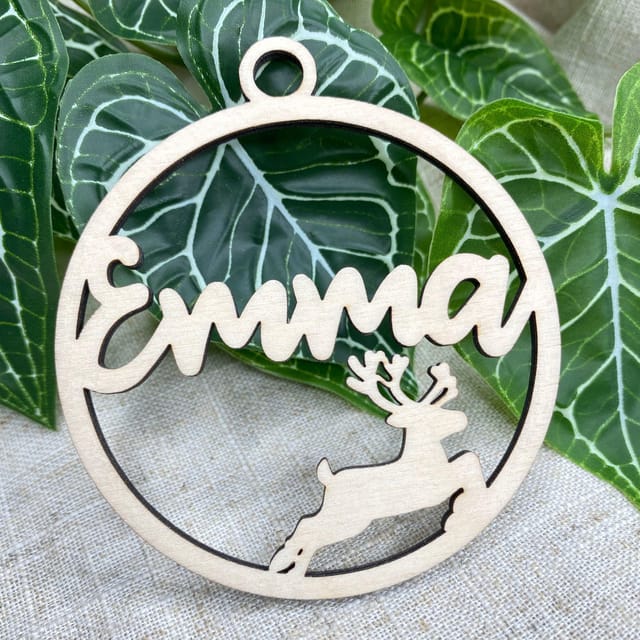 Könnte beinhalten: Ein kreisförmiges Ornament aus hellem Holz mit dem Namen Emma in Schreibschrift. Unter dem Namen befindet sich die ausgeschnittene Silhouette eines springenden Rentiers. Das Ornament hat oben eine kleine kreisförmige Aufhängung.