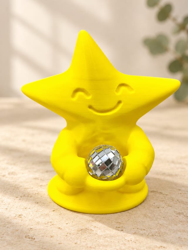 KI-bearbeitetes Produktbild: Gelbe Tanzfunke-Sternfigur mit lächelndem Gesicht und separater silberner Mini-Discokugel auf hellem Kalkstein vor cremefarbenem Hintergrund.