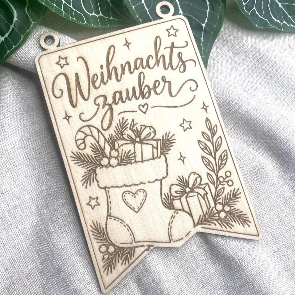 Ein rechteckiger Holzwimpel mit Gravur im Format A6 und zwei oberen Ösen liegt auf grauem Leinenstoff, mit dem Schriftzug Weihnachtszauber und Weihnachtsmotiven.