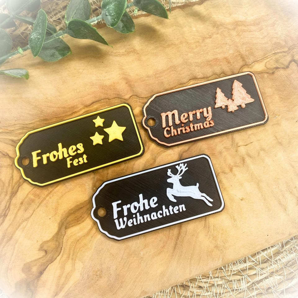 Könnte beinhalten: Drei Weihnachtsgeschenkanhänger mit schwarzem Hintergrund. Ein Anhänger hat den Text "Frohes Fest" mit gelben Sternen. Ein anderer sagt "Merry Christmas" mit zwei Bäumen. Der dritte Anhänger hat ein Rentier und den Text "Frohe Weihnachten."
