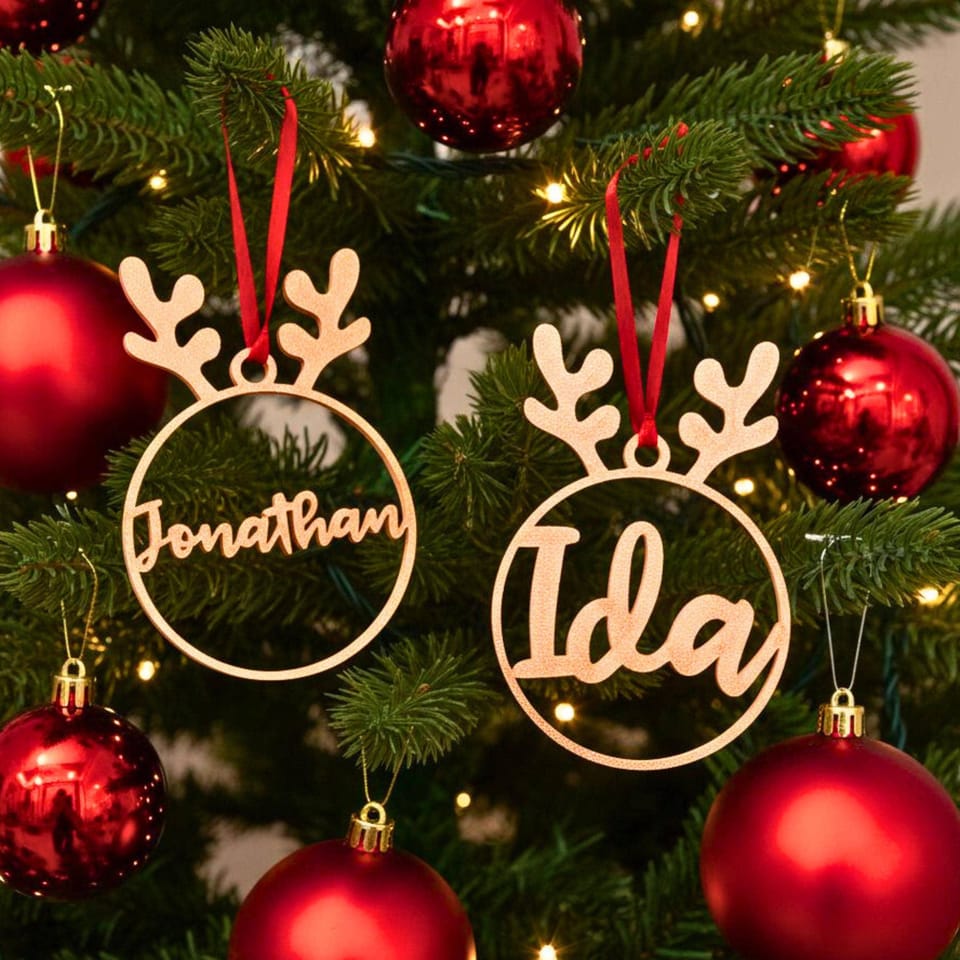 Zwei personalisierte Christbaumkugeln in Rentier-Form mit den Namen Jonathan und Ida hängen an roten Bändern an einem grünen Tannenbaum zwischen roten Kugeln.