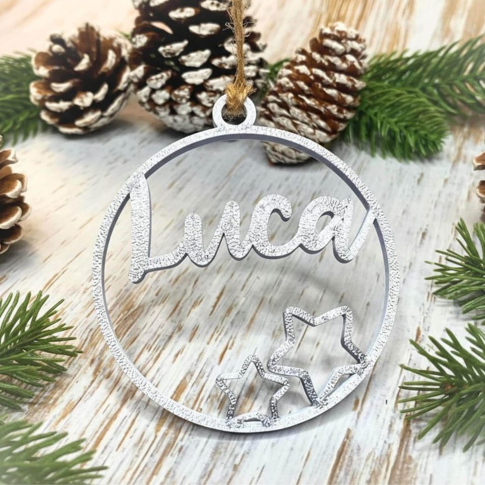 Ein silberner 3D-gedruckter Weihnachtsanhänger mit dem Namen Luca und zwei Sternen, hängend vor Tannenzapfen auf Holzgrund.