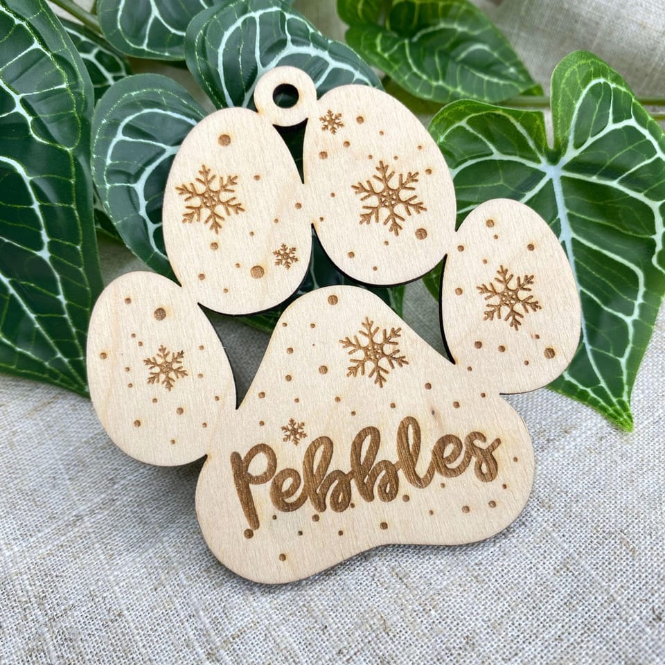 Könnte beinhalten: Ein helles Holzornament in Form eines Pfotenabdrucks mit drei ovalen Zehenballen und einem großen Hauptballen. Jeder Bereich ist mit Schneeflockenmustern und kleinen Punkten graviert. Der Name Pebbles ist in Schreibschrift eingraviert.