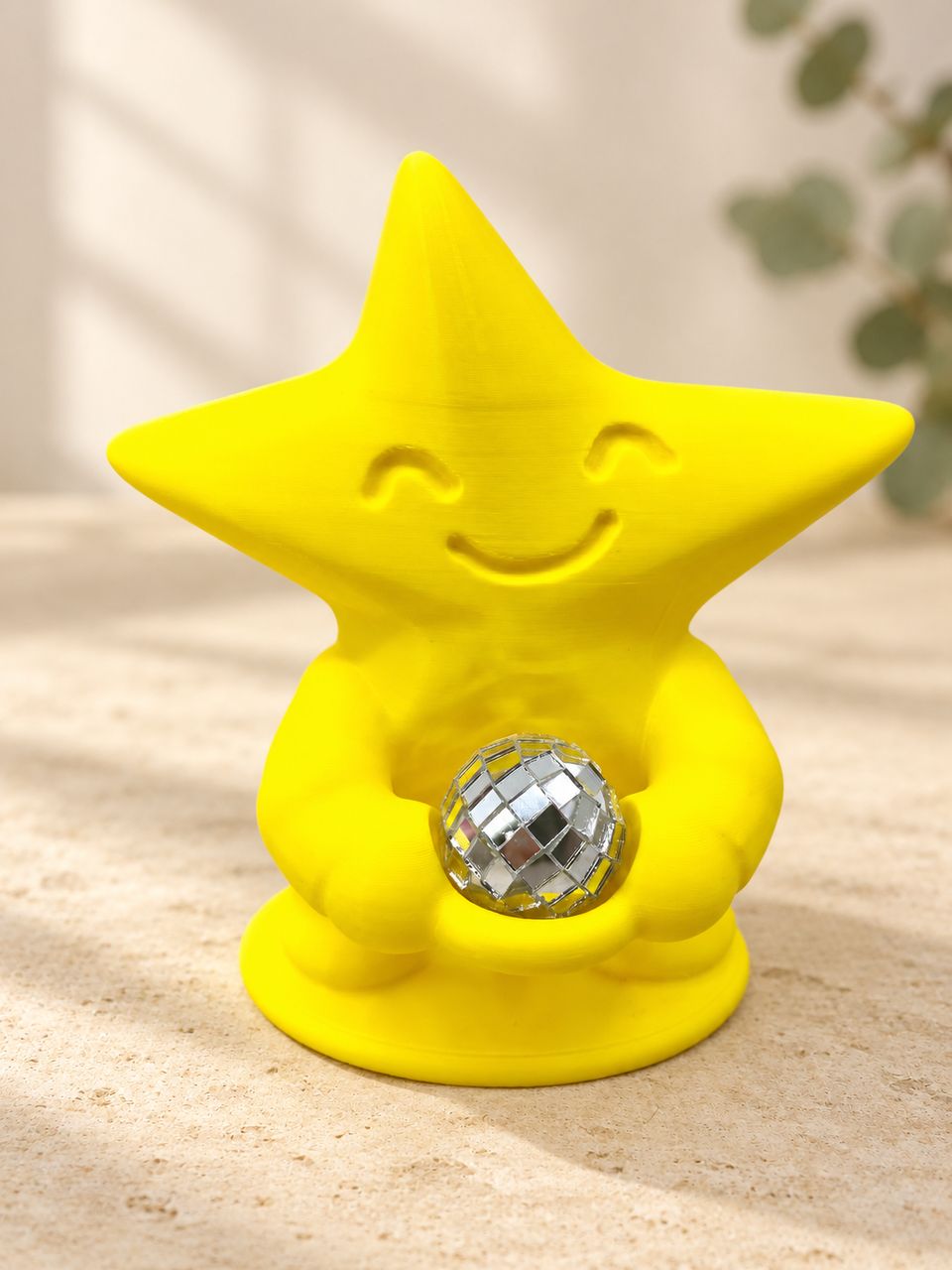 KI-bearbeitetes Produktbild: Gelbe Tanzfunke-Sternfigur mit lächelndem Gesicht und separater silberner Mini-Discokugel auf hellem Kalkstein vor cremefarbenem Hintergrund.